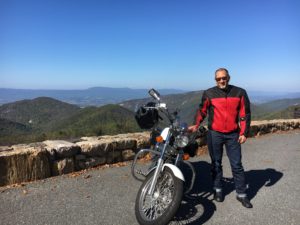 Man standing by motorcycle, scenic mountain view.